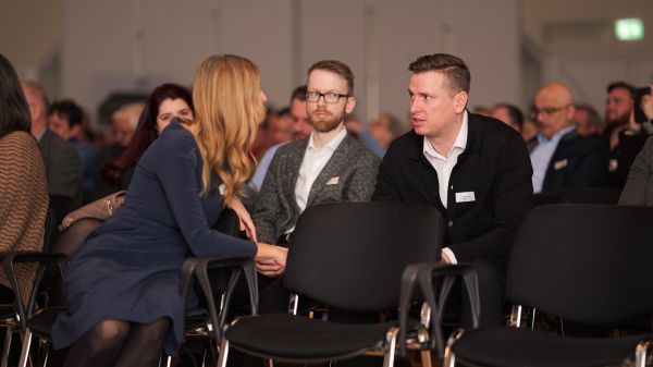 Eine Frau und ein Mann sitzen im Publikum und unterhalten sich. Dabei dreht sich die Frau nach hinten, um mit dem Mann zu sprechen.