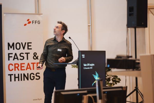 Ein Mann mit kurzen dunklen Haaren, grünem Shirt und schwarzen Hosen spricht ins Microphon.