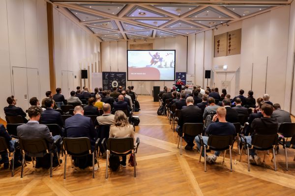 Der ganze Veranstaltngsraum von Hinten mit Blick auf die Bühne. Das Publikum sitzt in Stühlen.