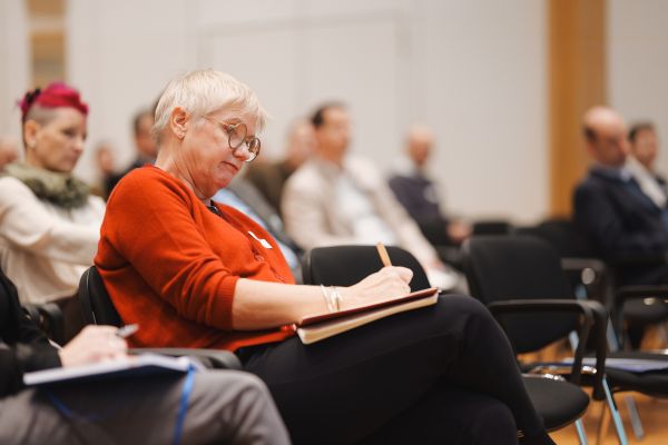 Frau mit kurzen weißen Haaren, rotem Pulli und schwarzer Hose sitzt im Publikum und schreibt mit.