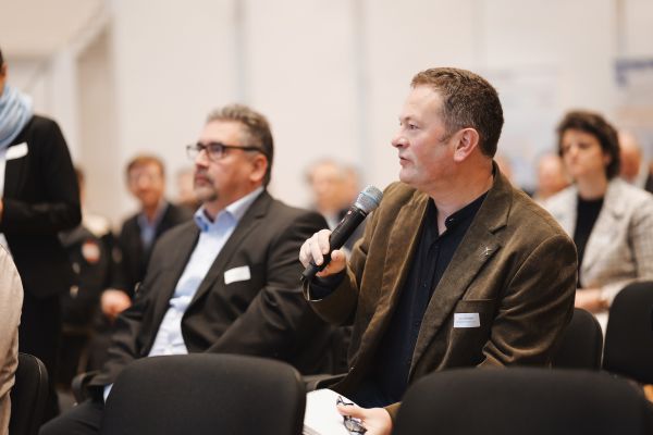 Zwei Männer im Publikum, einer spricht in ein Microphon.