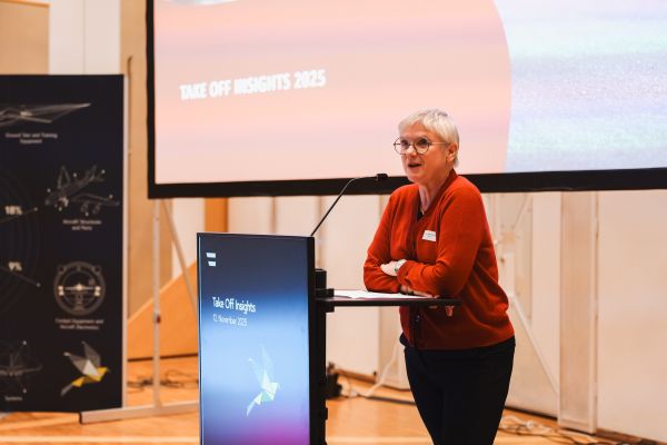 Frau mit kurzen weißen Haaren, rotem Pulli und schwarzer Hose spricht am Rednerpult.