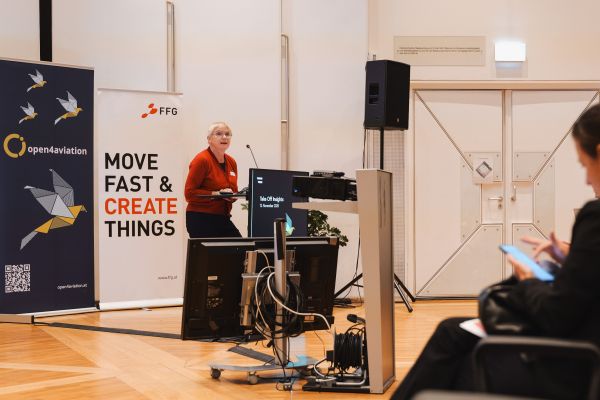 Frau mit kurzen weißen Haaren, rotem Pulli und schwarzer Hose spricht am Rednerpult.