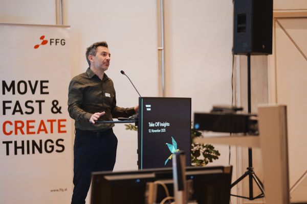 Ein Mann mit kurzen dunklen Haaren, grünem Shirt und schwarzen Hosen spricht ins Microphon.