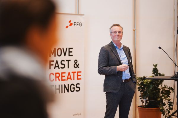 Ein Mann Mit Sakko und blauem Hemd steht beim Rednerpult.