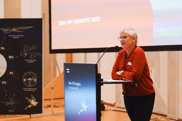 Frau mit kurzen weißen Haaren, rotem Pulli und schwarzer Hose spricht am Rednerpult.