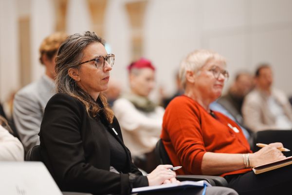 Frau mit kurzen weißen Haaren, rotem Pulli und schwarzer Hose sitzt im Publikum und schreibt mit.